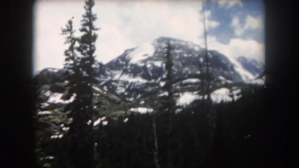 1962 Summit County, Colorado 8mm frame showing a snow-capped mountain peak beyond evergreen trees and shadowed forest.