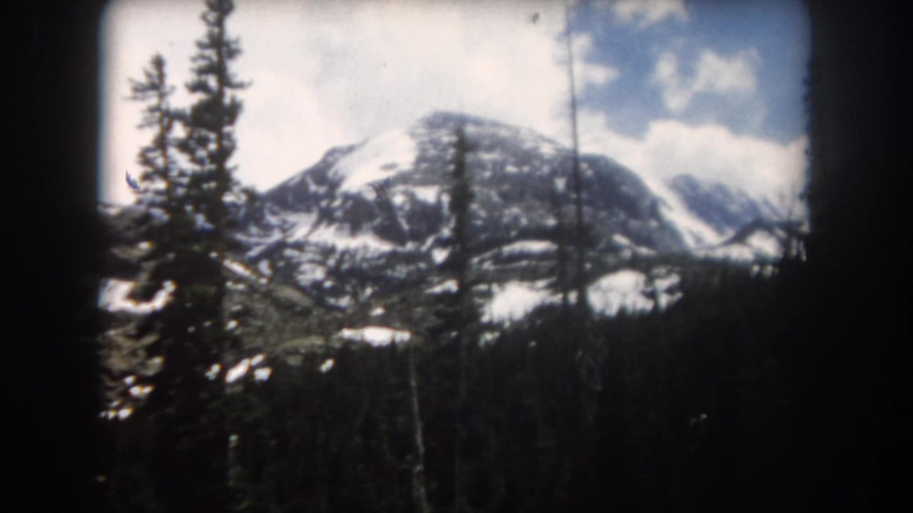Evergreens frame a snowy mountain ridge under a partly cloudy sky.