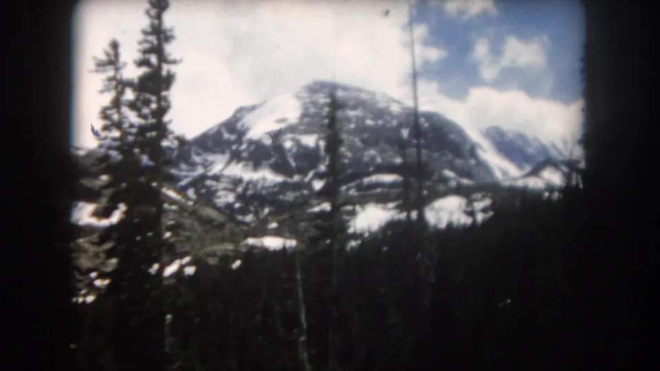 1962 Summit County, Colorado home-movie frame with evergreens in front of a snow-streaked mountain ridge and pale clouds.