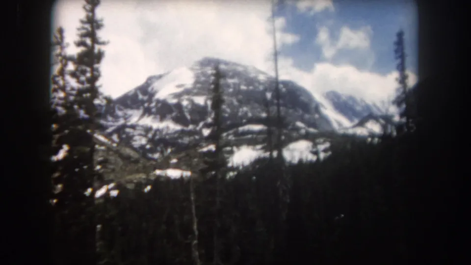 1962 Summit County, Colorado 8mm frame showing a snowy peak partly screened by tall evergreen and bare tree trunks.