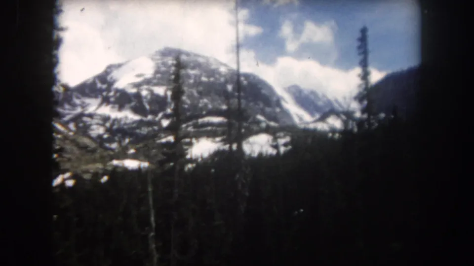 1962 Summit County, Colorado 8mm frame showing snowy mountains behind a dense evergreen forest and scattered clouds.