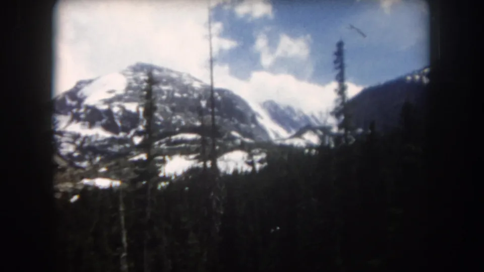 1962 Colorado 8mm still showing a snow-covered mountain slope above a shaded pine forest and bright clouded sky.