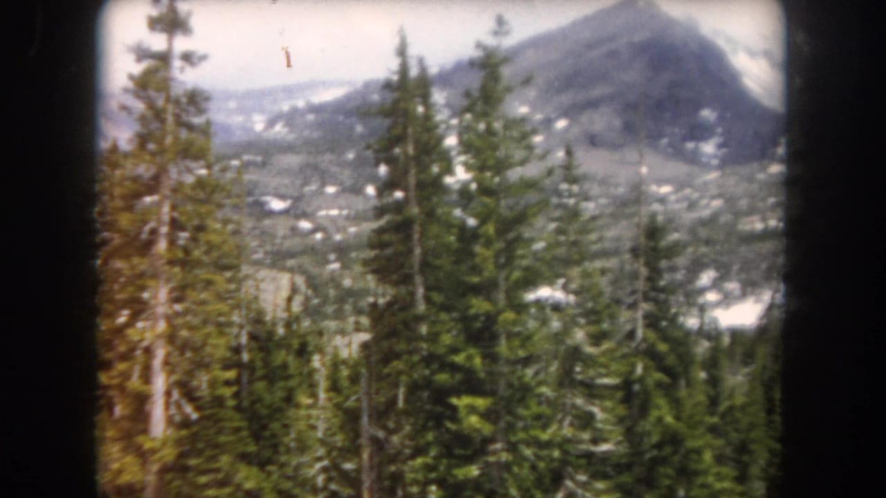 1962 Summit County, Colorado frame with tall conifers in the foreground and a broad snow-patched mountain slope beyond.