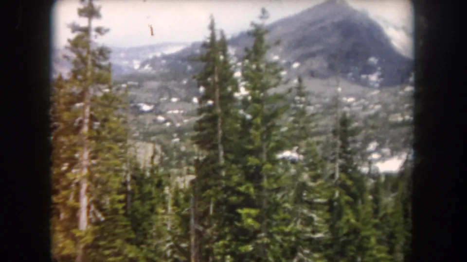 1962 Summit County, Colorado frame with tall conifers in the foreground and a broad snow-patched mountain slope beyond.