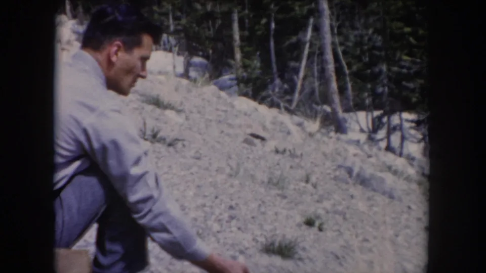 In the 1962 Colorado home-movie footage, the man crouches and reaches toward the rocky ground to lure the chipmunk.