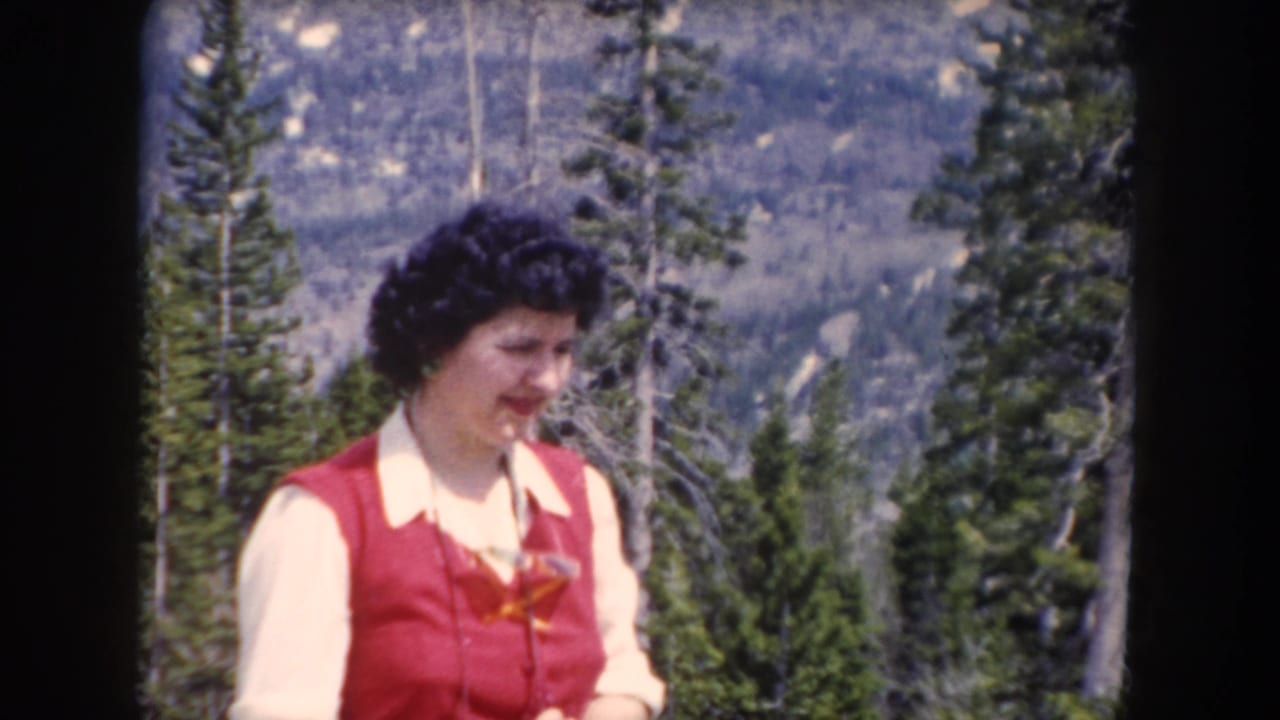 Woman in a red vest standing before evergreen trees and mountain slopes.