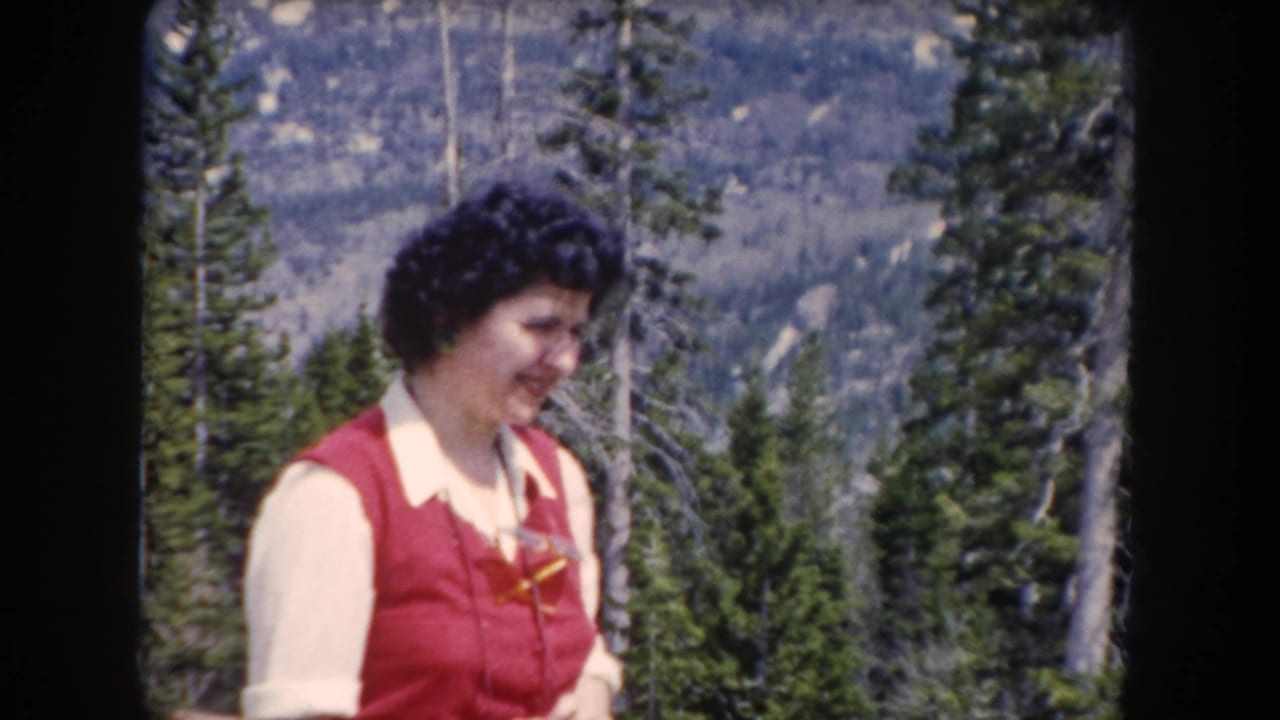Woman in red vest looking down with pine trees behind her.