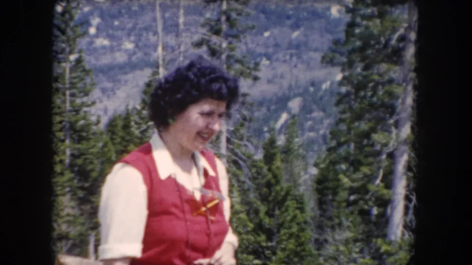 Smiling adult woman in red vest appears in a Summit County, Colorado forest scene from 1962 home-movie footage.