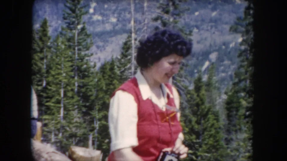 Adult woman in red vest holds a small object, possibly a camera, in a 1962 Summit County mountain forest home-movie frame.