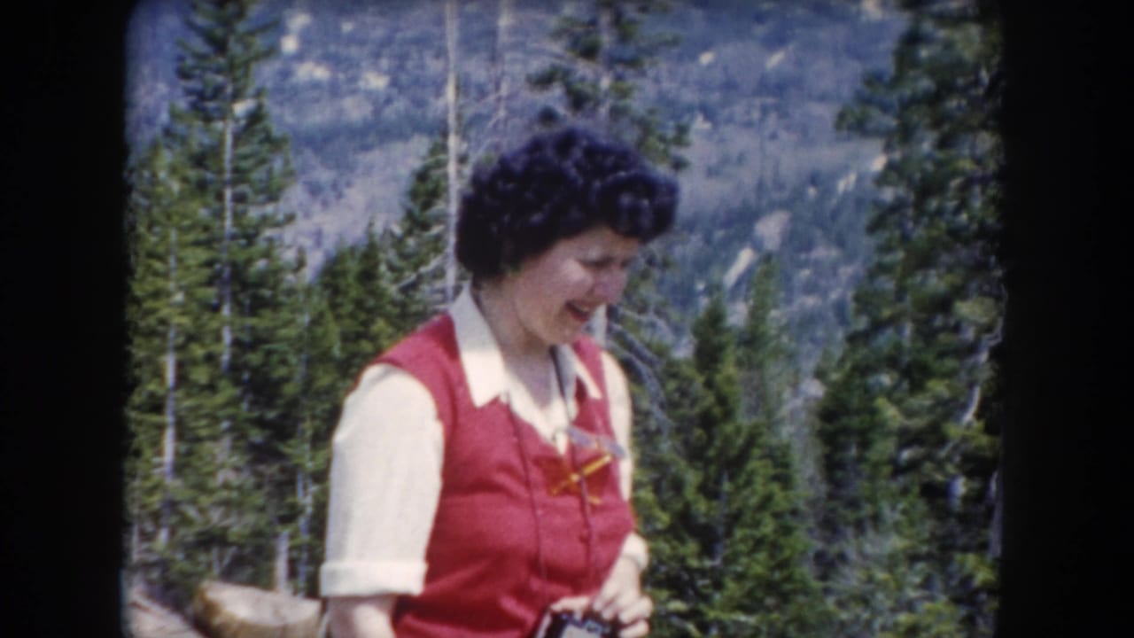 In 1962 Summit County, Colorado footage, a woman in a red vest and white collared shirt stands in a pine forest with mountains behind her.