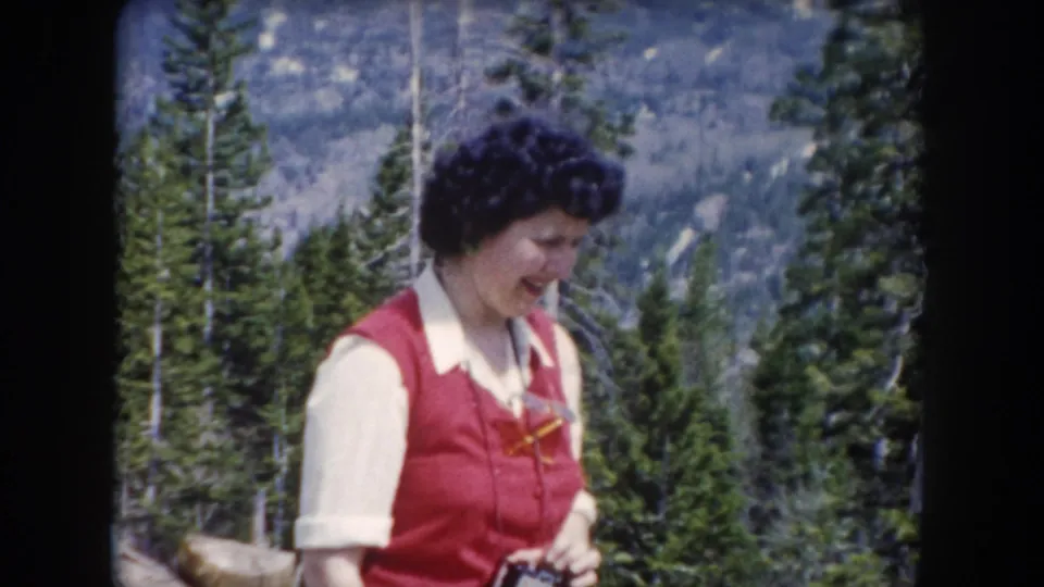 In 1962 Summit County, Colorado footage, a woman in a red vest and white collared shirt stands in a pine forest with mountains behind her.