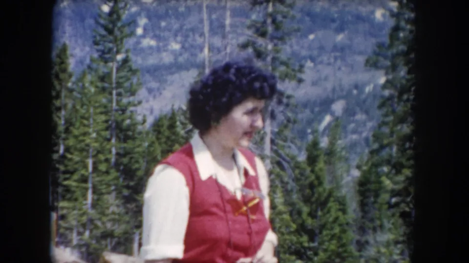 A woman in a red vest appears mid-reaction in a Summit County, Colorado forest, with evergreen trees and a mountain slope visible behind her.