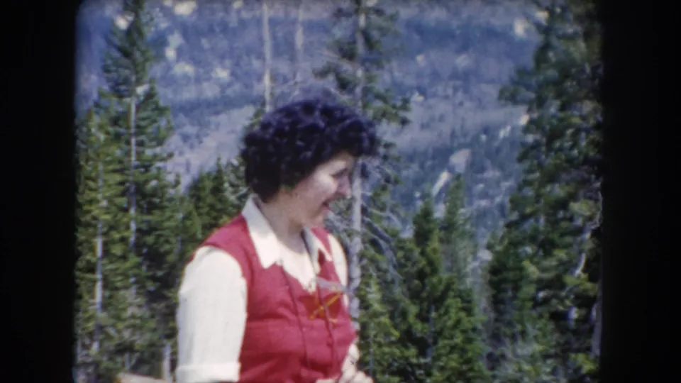 The woman smiles in the 1962 mountain forest footage, wearing a red vest and white collared shirt with a camera partly visible near her hands.