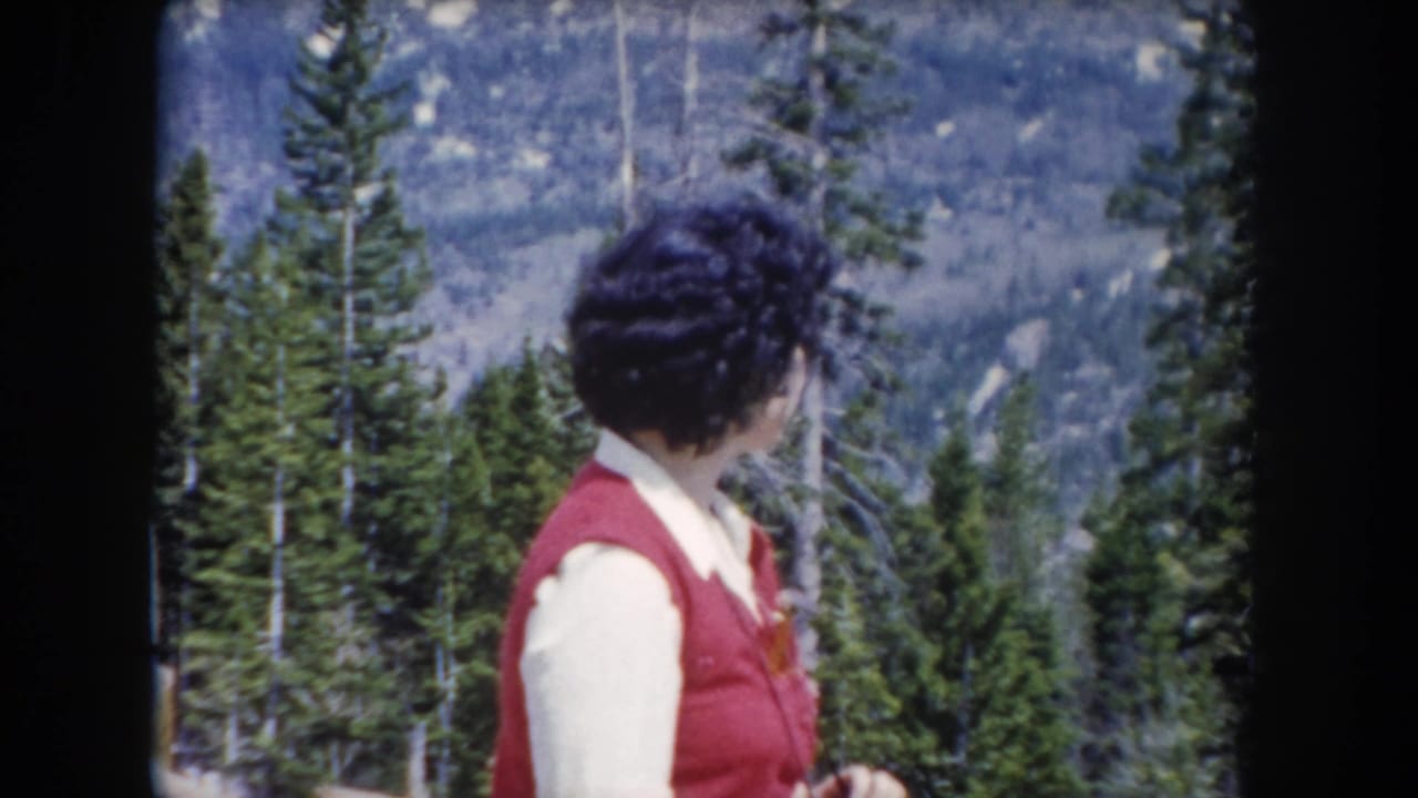 In 1962 Summit County, Colorado footage, a woman in a red vest faces away from the camera in a mountain pine forest.