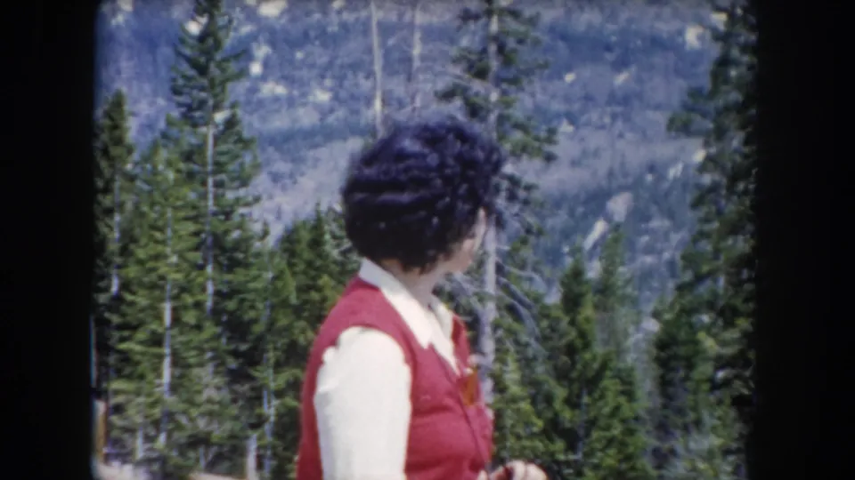 In 1962 Summit County, Colorado footage, a woman in a red vest faces away from the camera in a mountain pine forest.