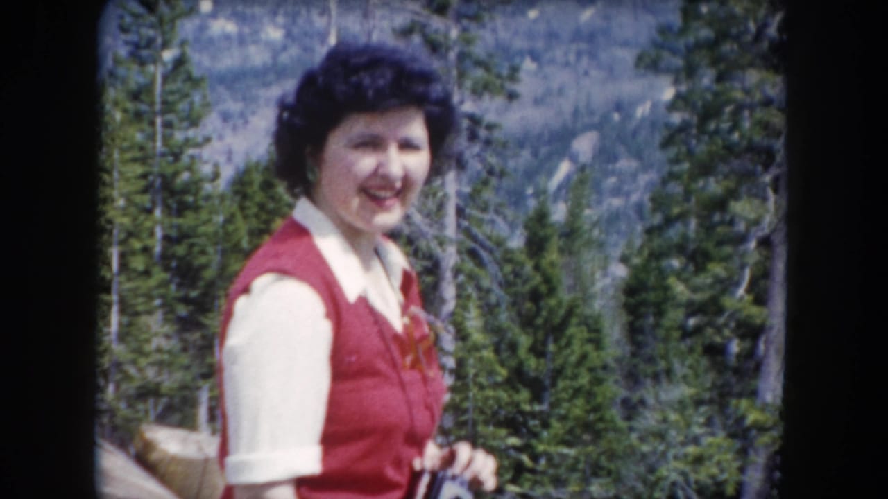 The woman smiles toward the camera while standing among pines in 1962 Summit County, Colorado home-movie footage.