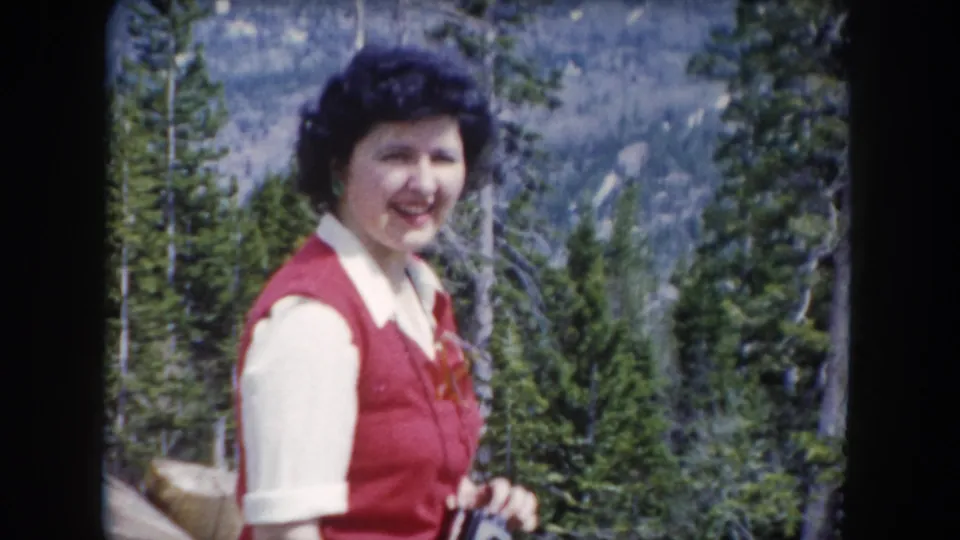 The woman smiles toward the camera while standing among pines in 1962 Summit County, Colorado home-movie footage.