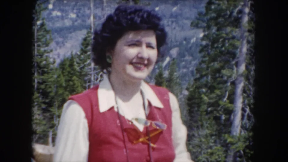 A smiling woman in a red vest and white blouse holds a small camera against a pine-covered mountain backdrop in Colorado.