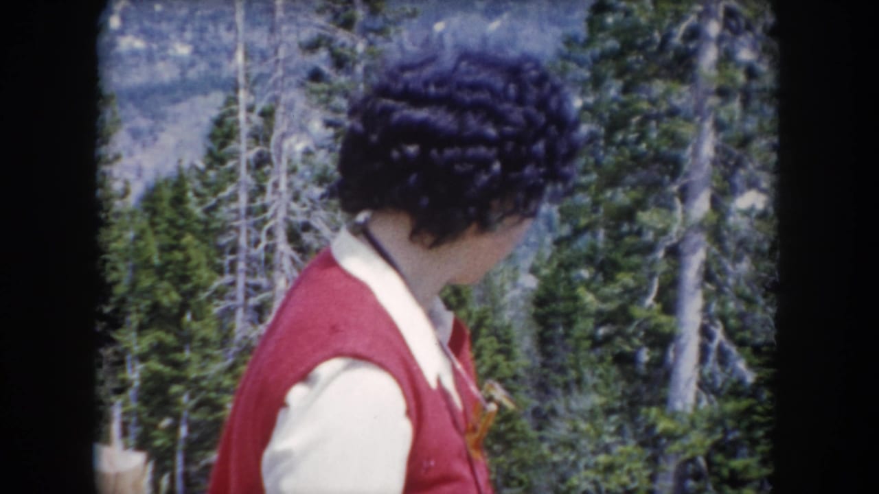 In 1962 Summit County, Colorado footage, a woman in a red vest turns her head beside evergreen trees and mountain terrain.