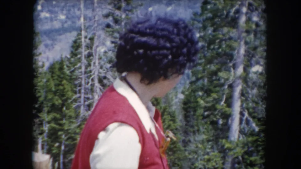 In 1962 Summit County, Colorado footage, a woman in a red vest turns her head beside evergreen trees and mountain terrain.