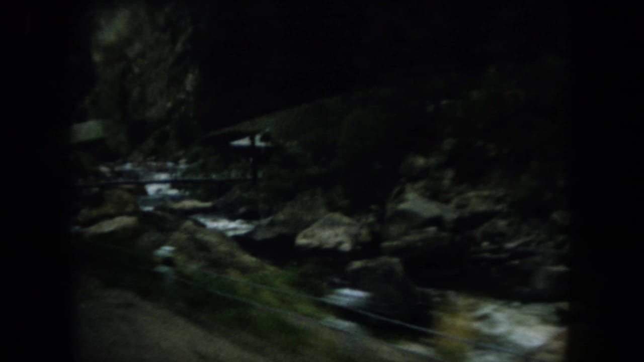 Rocky creek in a wooded area with a small bridge or rail in the distance.