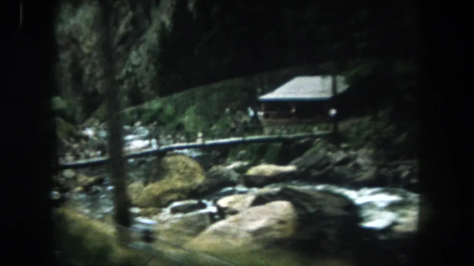 1962 Summit County frame showing a small streamside cabin, boulders, water, and dense wooded slopes.