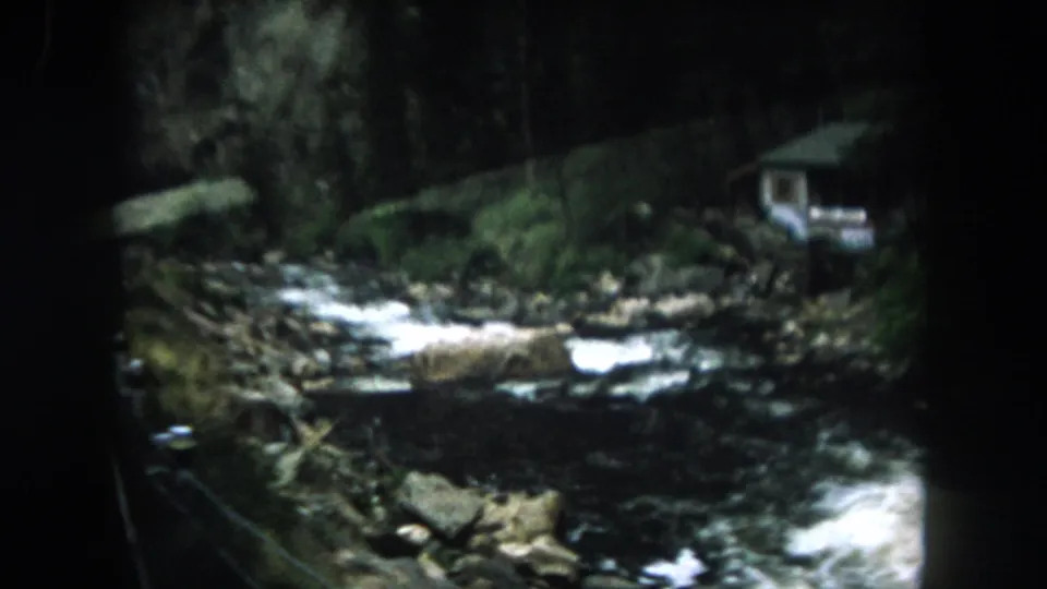 1962 Summit County 8mm frame showing white water over rocks, wooded creek banks, and a small structure at right.