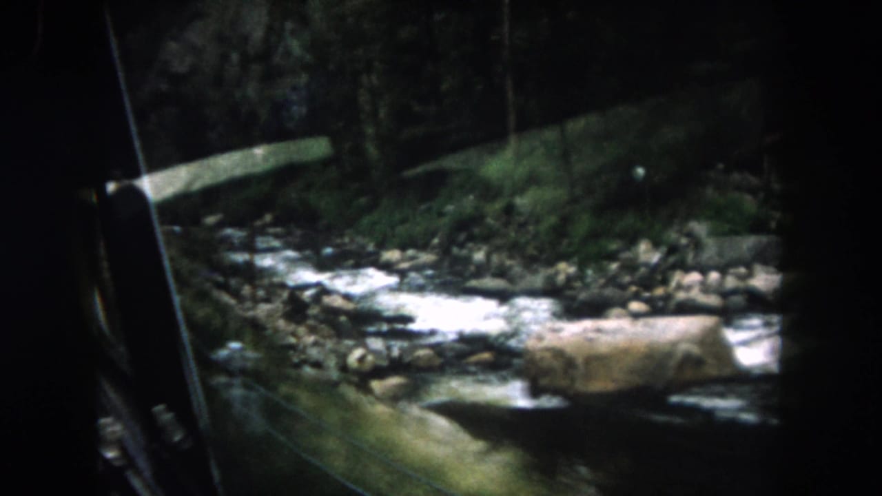 1962 8mm view in Summit County, Colorado, looking from a moving vehicle toward a rocky river, forested bank, and roadside rail.