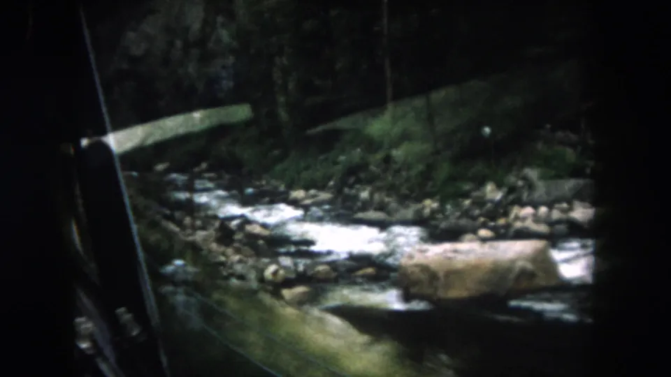 1962 8mm view in Summit County, Colorado, looking from a moving vehicle toward a rocky river, forested bank, and roadside rail.