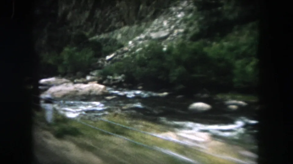 Summit County mountain stream flows over pale rocks beneath dense trees in soft 8mm color footage.