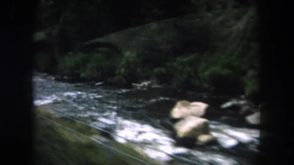 Vehicle-shot 8mm view of a rocky river beside a wire fence and forested bank in Summit County.