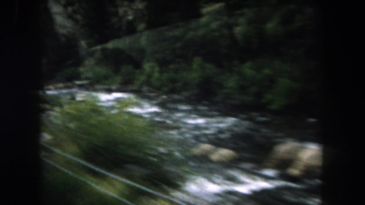 1962 Summit County, Colorado home-movie frame showing a rocky river and forested bank from a moving vehicle.