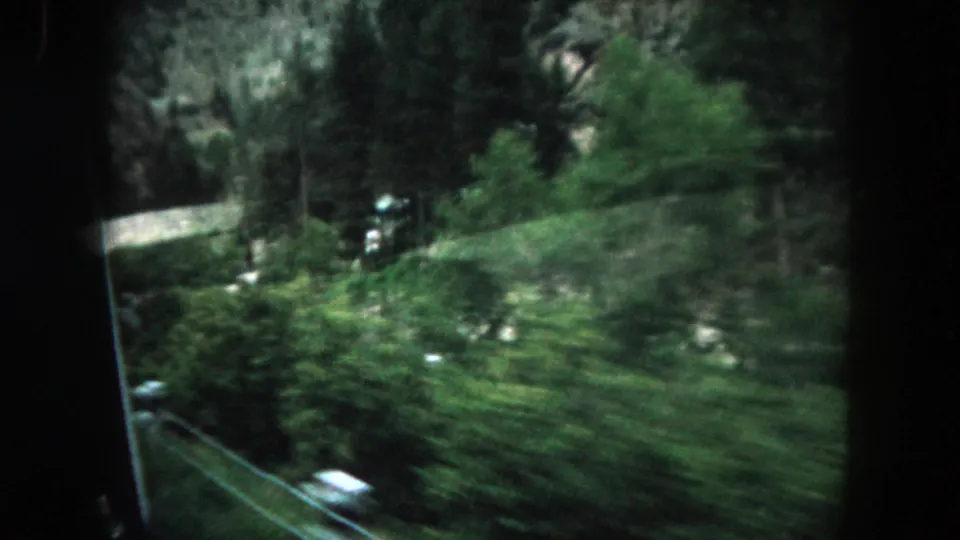 1962 Summit County, Colorado frame of a forested roadside and rocky slope seen from a moving vehicle.