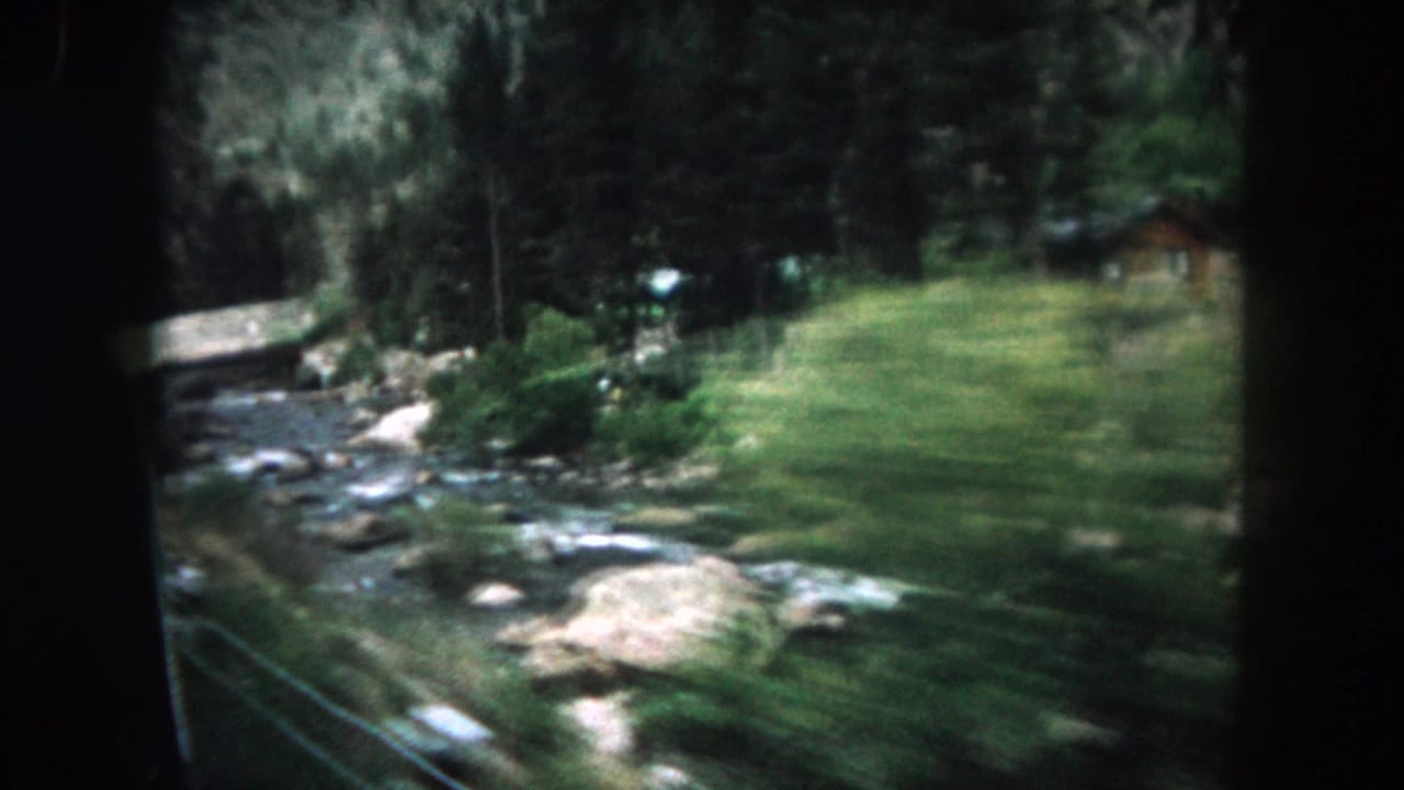 1962 Summit County car-window view of a rocky mountain river, grassy bank, evergreens, and a small distant structure.