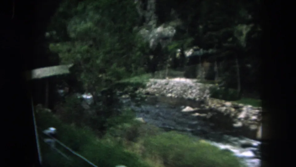 The car passes a shaded Summit County river bend with boulders, dense trees, and a roadside rail partly visible at frame edge.