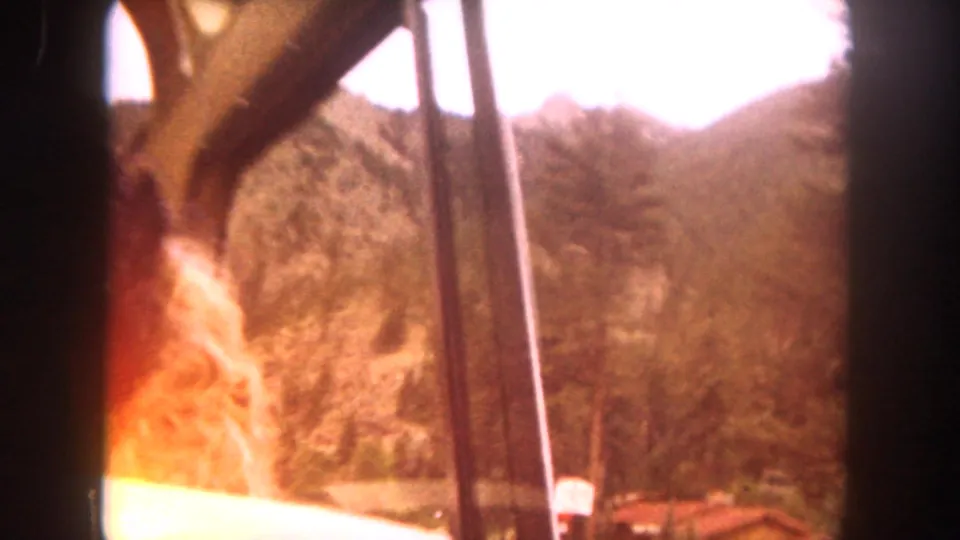 Warmly exposed 1962 home-movie frame from inside a car, showing a partial passenger figure, window posts, forested hillside, and red-roofed structure.
