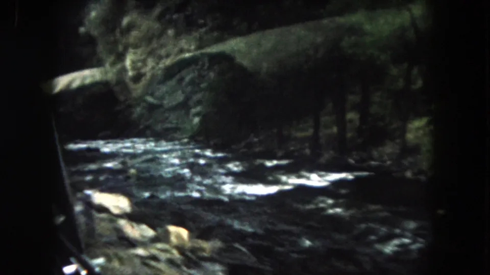 1962 Colorado passenger footage looking across a fast river toward trees and rocky banks along a mountain road.