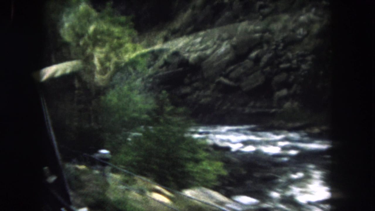 1962 Summit County, Colorado 8mm frame looking from a car window toward a rushing stream, rocks, and dark evergreens.