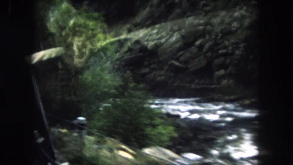 1962 Summit County, Colorado 8mm frame looking from a car window toward a rushing stream, rocks, and dark evergreens.