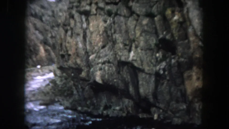 Summit County, Colorado home-movie frame showing a rock wall, moving water, and dark foreground branches near the road.