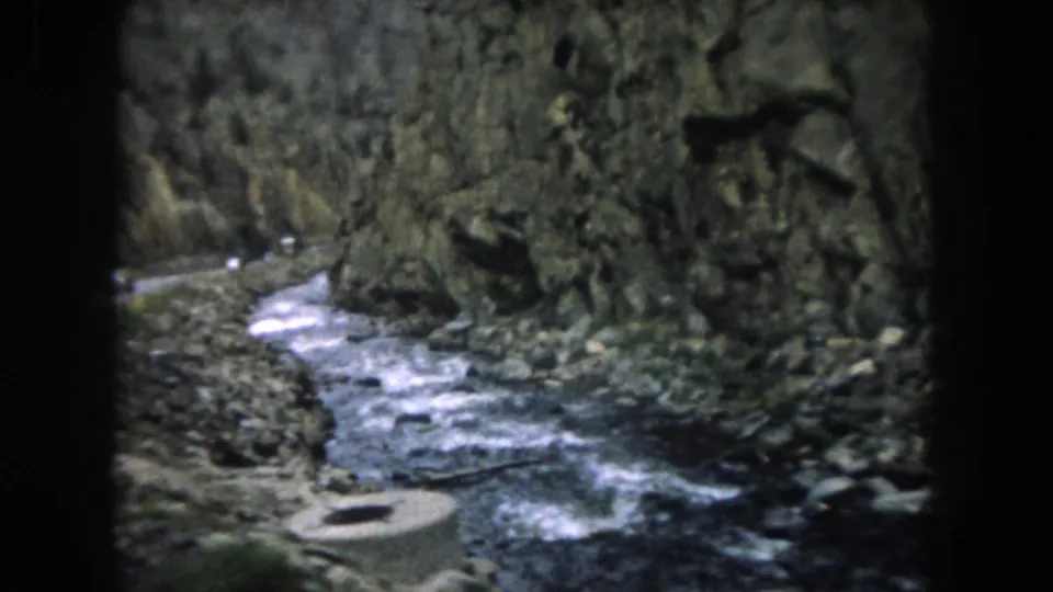 1962 Summit County, Colorado 8mm frame with a rocky stream, cliff wall, roadside stones, and vehicle-window obstruction.