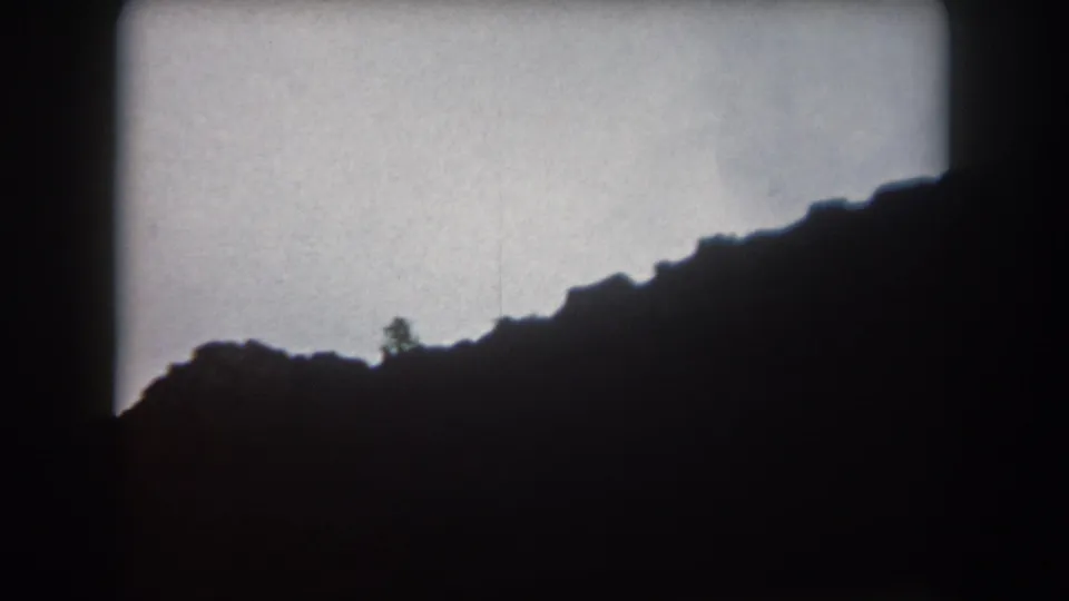 1962 Colorado home-movie frame with a dark mountain ridge sloping upward below a washed, overcast dusk sky.