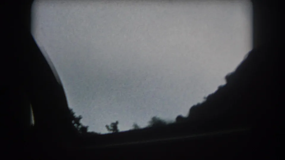 Summit County road-view frame with a dark hillside, sparse tree silhouettes, and sky from inside the moving car.