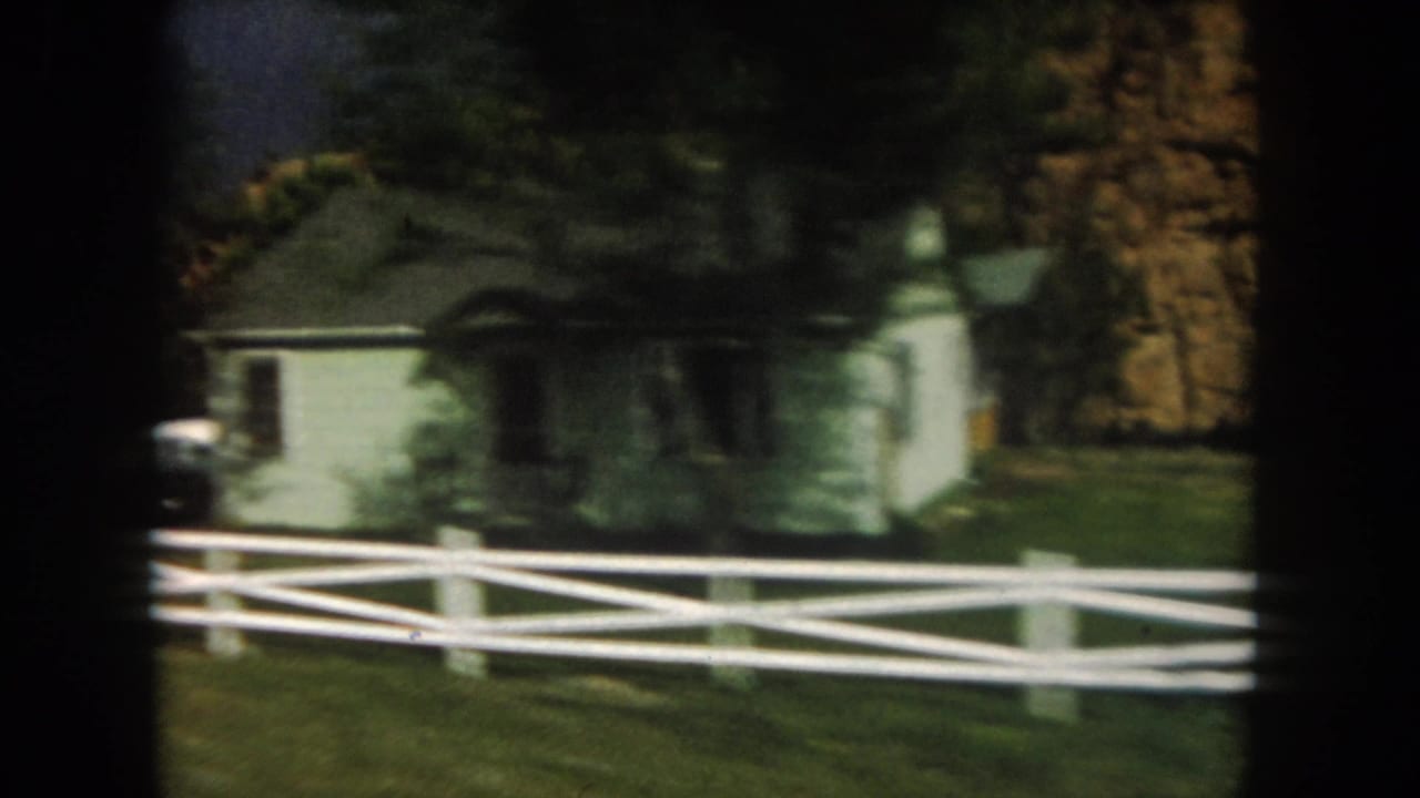 White farmhouse behind a white rail fence with trees and grass.