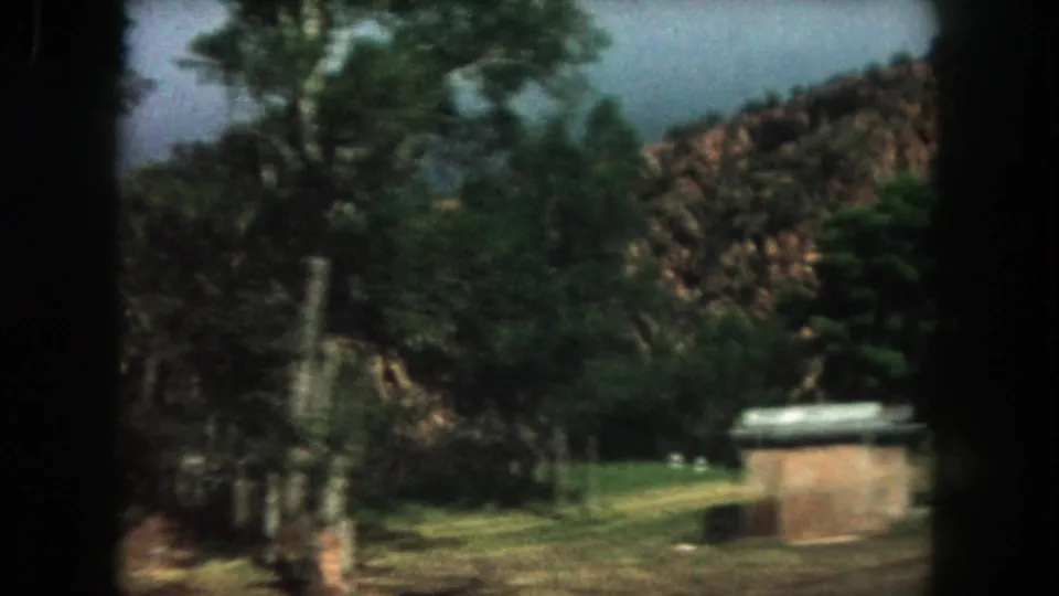 1962 Summit County, Colorado home-movie frame showing a small brick structure, open grass, trees, and rocky hillside.