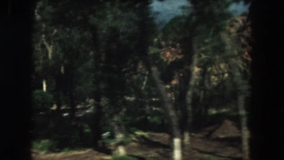 1962 Summit County, Colorado home-movie frame with shaded trees, grass, fence elements, and a rocky hillside beyond.