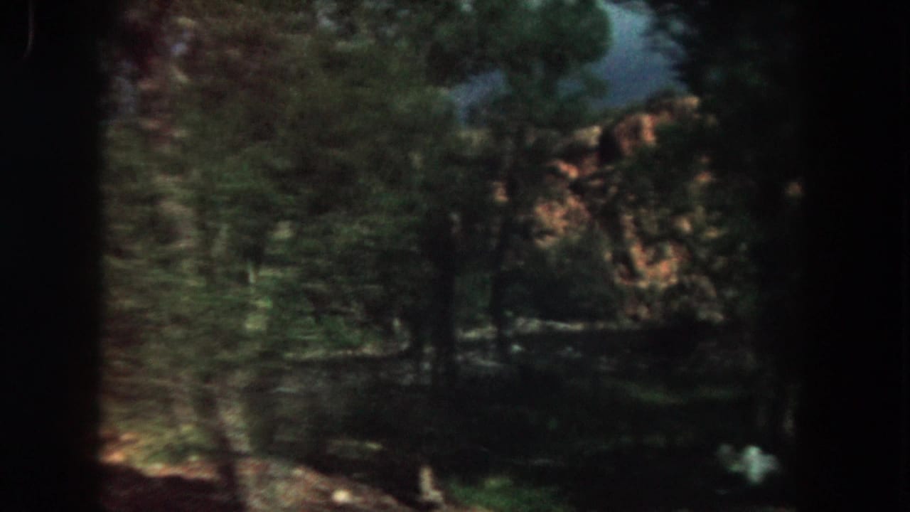 1962 Summit County, Colorado 8mm frame showing dense trees, rocky hillside, and dark gate shadows during a scenic drive.
