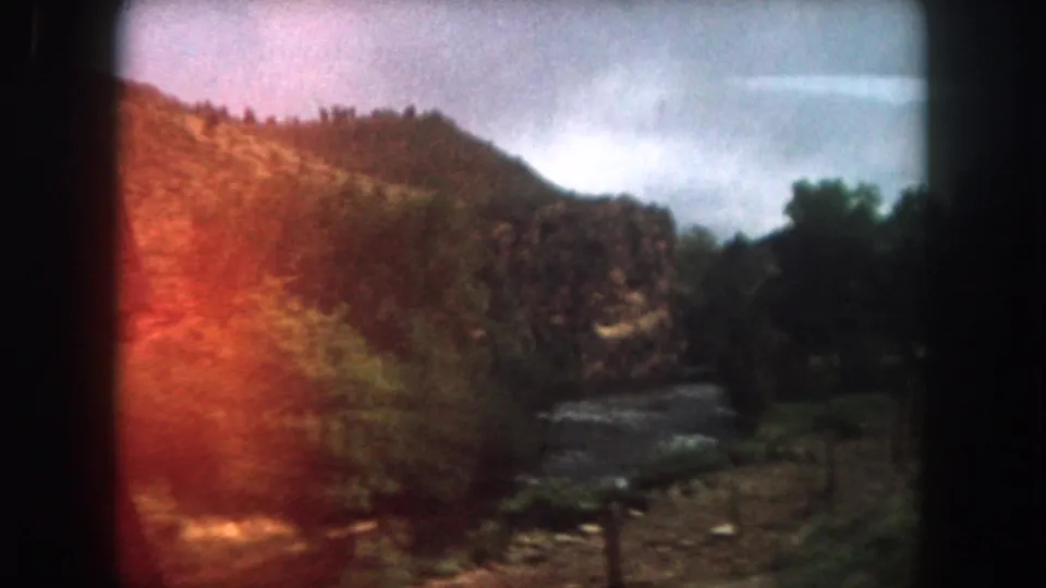 Summit County river valley in 1962 home-movie footage, with forested hills, cloudy sky, roadside fence posts, and red film flare.