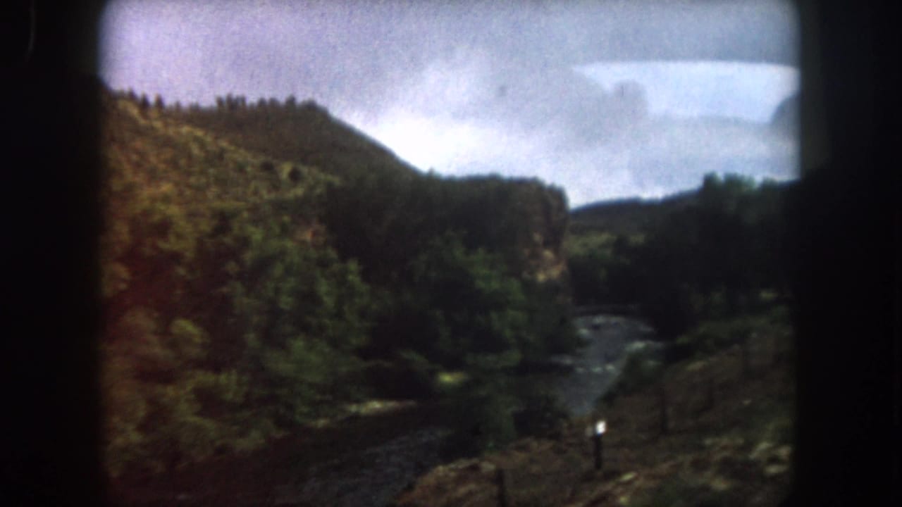 1962 Summit County, Colorado home-movie frame showing a river valley, wooded hillside, roadside fence, and dark car-window edges.