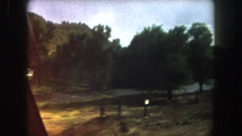 1962 Summit County, Colorado frame showing sunlit roadside ground, fence rails, dense trees, river water, and mountain slope.